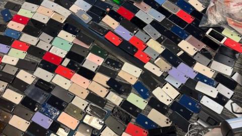 A large collection of smartphones spread across a grey carpeted floor in an office setting. The phones are arranged in a dense, irregular grid, with their backs facing upward. They vary in colour, including black, white, silver, gold, red, blue, green, and purple. Some phones appear scratched or damaged, while others look intact. In the background is a clear plastic bin bag.