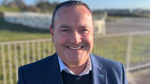 Head and shoulders image of John Pye smiling in a garden by the coast in the sunshine. He has short dark hair combed back and is wearing a white shirt under a dark round neck jumper and blue suit jacket