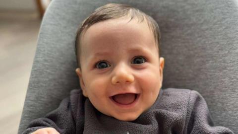 A baby boy is smiling with his mouth wide open. He is wearing a grey top and is sitting in a grey bouncer chair.