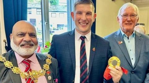 Gregg Stevenson holds his medal as he stands between the Mayor of Pendle, councillor Mohammad Aslam, who is wearing his ceremonial chain, and councillor Kevin Salter. All three men are smartly dressed and smiling.