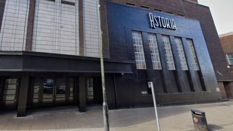 The venue has a blue brick and glass entrance on a main street in the city, with a large sign that reads Astoria.