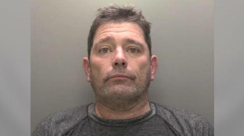 A police custody picture showing the head and shoulders of a man with short brown hair and stubble wearing a grey T-shirt against a grey backdrop. 