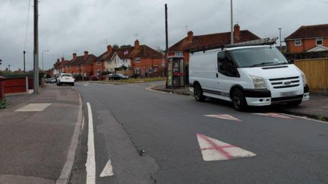 A residential street on a cloudy day with a white van parked on the right hand side and a white car parked on the left, a little further away. On the road, the white triangle indicating the presence of a speed bump has a red cross spray painted on it.