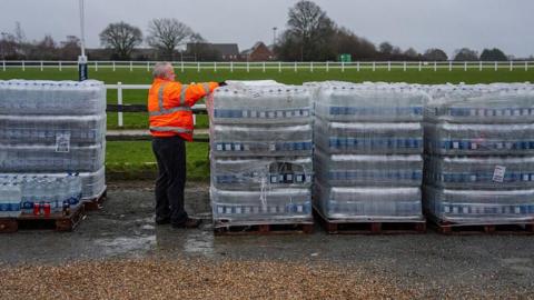 A man in an orange high vis coat next to creates of bottled water.