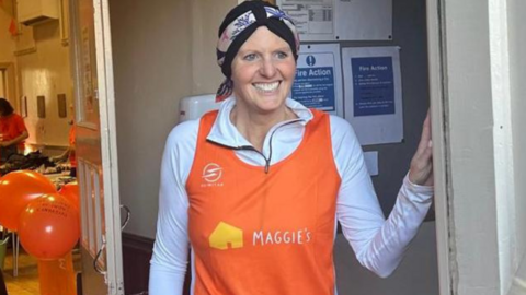 A woman stood in a doorway and smiling wearing an orange top that says 'Maggie's'