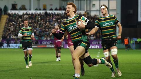George Hendy, wearing the green, gold and black of the Northampton Saints, celebrates in the act of scoring a try against the Sale Sharks at Franklin's Gardens. Various teammates can be seen in the background.
