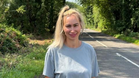 A woman in a light blue T-shirt is standing on the side of a country lane. She has blonde hair tied in a ponytail that falls onto her right shoulder. Long green grass, trees, and bushes surround her on the verges.