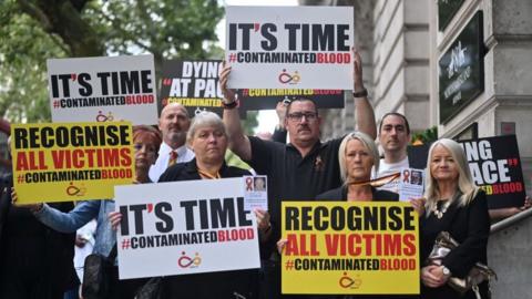 Demonstrators outside the public inquiry in 2023 hold placards related to the infected blood scandal reading: "It's time" and "recognise all victims". 