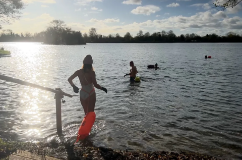 A group of swimmers walk into South Cerney lake on a clear bright winter's day. The sun is reflecting off the water and the trees that border the lake can be seen in the distance
