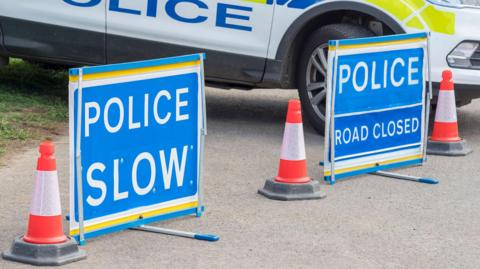 A stock image of blue police "road closed" signs, orange traffic cones and a police car in the background.