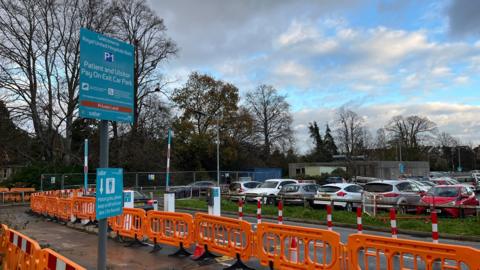 The P1 car park at the Royal United Hospital in Bath. The image shows two blue signs for the patient and visitor car park. There are orange barriers in the foreground and several rows of cars are parked behind a grass verge.
