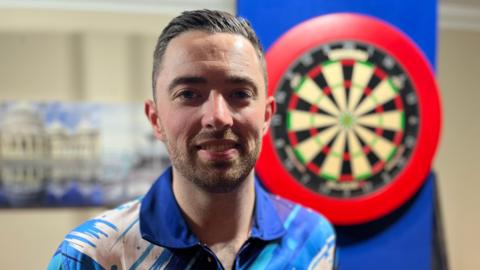 Luke Humphries in his blue darts t shirt with short brown hair and a dart board behind him.