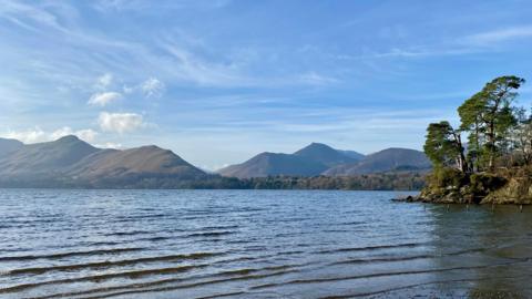 Large lake looking out to mountains with trees to the right under a blue sky with wispy cloud