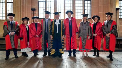 Nine people stand facing the camera as they pose for a picture ahead of an honorary degree ceremony at the University of Oxford. They are in a variety of honorary robes and caps. From left, a man with glasses wears a red silk robe with grey and graduation cap, followed by to two ladies wearing red silk robes and doctoral Tudor bonnets, and a man with glasses wearing a red silk robe with grey and graduation cap. In the middle stands a man with a dark suit, white shirt and gold and black robe and graduation cap with gold tassel. To the right of him, a man in a red robe and Tudor bonnet, a woman in a red and grey robe with graduation cap, a woman in a red robe and Tudor bonnet and a woman in a red and grey robe and graduation cap.
