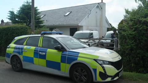 A marked police car is parked outside a rural house, with a gate and hedges partially blocking the driveway. An officer stands near two vans inside the property, suggesting an active police presence at the scene.