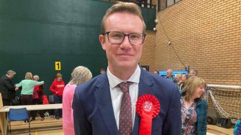 Tim Roca standing in a community sports hall after being elected Labour MP. He is wearing glasses, a navy blue suit and red tie with white spots, and has a red Labour rosette pinned to his lapel.