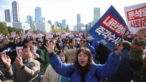 A group of men and women cheer and hold placards outside the national assembly in Seoul on Wednesday, with the Seoul skyline visible in the background.