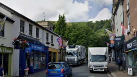 Two lorries can be seen coming down the street in Malvern with a car going in the opposite direction. There are shops on either side of the vehicles.