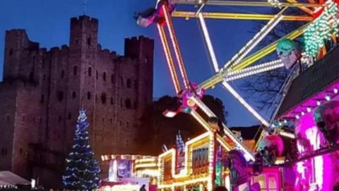 A festive scene of lit funfair rides in foreground with a lit Christmas tree and Rochester castle in background.