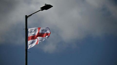 A St George's flag attached to a lampost is blowing in the wind against a slightly grey looking sky. 