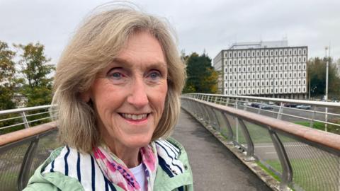 Maureen Millar pictured at a bridge leading to Marlborough House. She has shoulder length light hair and is wearing a green coat with a pink patterned scarf. She is smiling.