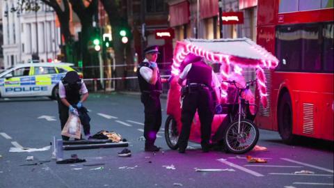 Police officers attending the scene of the crash at Charing Cross. A pink pedicab with lights wrapped around the frame appears damaged and is next to a red London bus. Debris is strewn across the road near the officers' feet and cordon tape is visible in the blurred background.