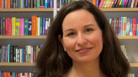 Iryna Bakaliar is looking at the camera and is standing in front of bookshelves full of Ukrainian books.