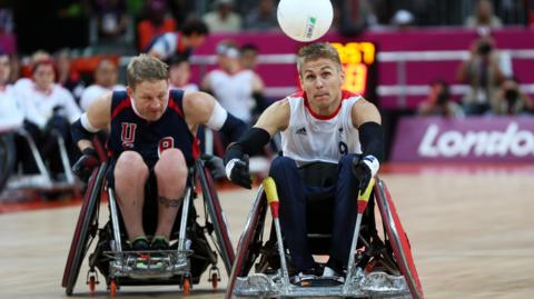 Wheelchair rugby players compete for the ball during a match at London 2012, with Steve Brown in a white Great Britain shirt and another in a dark USA kit racing side by side in specialised sports wheelchairs.