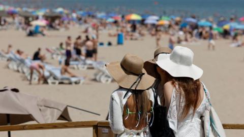 Three women with beach hats stand looking at a beach that has people sunbathing, sat under parasols, and swimming in the sea.