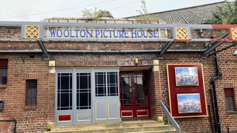 Front exterior of the one-storey brick cinema. Blue art deco sign saying Woolton Picture House is above the 1920s type doors.
