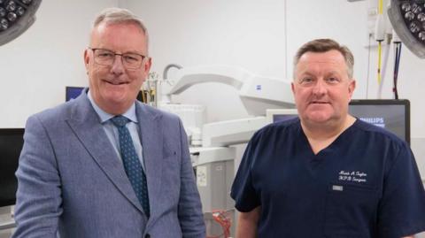 Two men stand in a surgical ward with surgical lights and machines. The man on the left is wearing a light blue suit, light blue shirt and teal tie. He has white hair, is wearing glasses and is smiling. The man on the right has brown and white hair and is wearing dark blue medical scrubs.