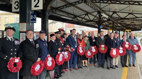 A number of people in military uniforms are holding wreaths and are gathered on a train platform. 