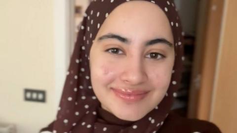 Anaba Khan, who is wearing a brown headscarf with white dots, smiling