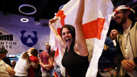A woman in a black top cheers and holds a flag of St George aloft as she celebrates England beating Spain in the final of the European Championships at Ashton Gate in Bristol