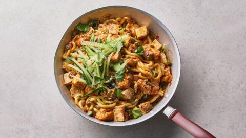 A pot with golden thick noodles and tofu topped with sliced cucumber