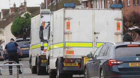 An army bomb expert dressed in black and wearing a back beret and blue bullet-proof vest looks at something on the ground on a Belfast street. Two white army vans are parked beside him