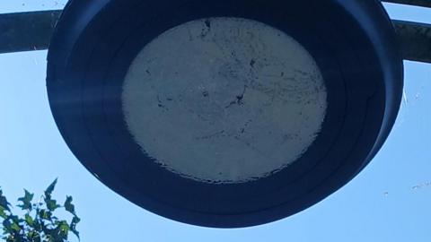 Close-up image taken from below of a dark circular-shaped street light. Its surface is scratched and marked. It appears to be in the centre of an arch, although only the immediate supporting structure of two metal bars, extending either side of the lamp, can be seen.