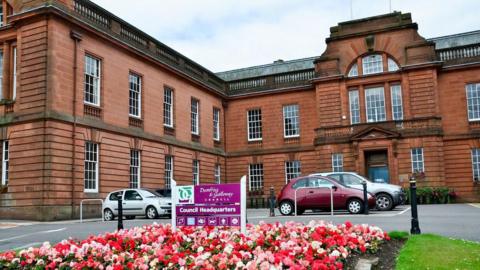 Dumfries and Galloway Council headquarters with flowers outside, it is a sandstone building with cars parked in front of it