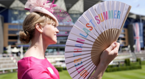 A woman in hot pink dress and pastel pink fascinator fans herself with a white fan decorated with the writing Hotter Then Last Summer while waiting on a grassy enclosure at Ascot Racecourse