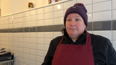 Fishmonger Angela Preece stands in her shop in front of a wall of white tiles, wearing a red apron, black fleece and purple hat. Weighing scales are visible in the background to the left