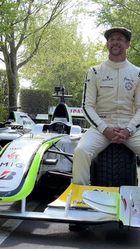 Jensen Button wearing wite racing overalls, sitting on the front wheel of a racing car.