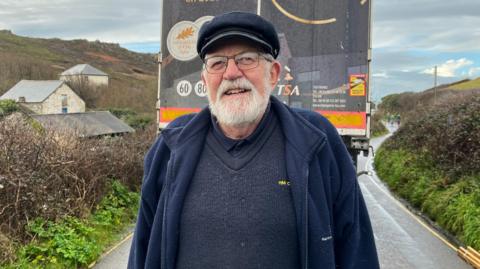 Barry Mundy is standing in front of the rear of the lorry. He is wearing a navy fishing cap and a navy fleece over a thick knitted navy jumper and navy shirt. He is smiling. He has a white beard and is wearing a pair of metal rimmed glasses.
