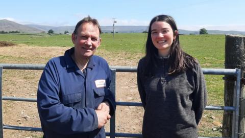 Andrew and Katie Laidlow in front of a farm gate with a field behind them and the fells visible on the horizon. Mr Laidlow has short brown hair and is wearing blue overalls. Miss Laidlow has shoulder-length dark hair and is wearing a grey fleece. Both are smiling at the camera.