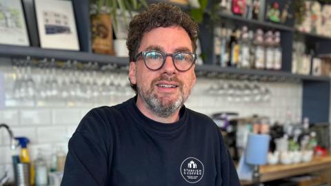 Tony Marks, owner of the Starfish and Coffee cafe in Brighton in the UK, stood in his cafe. He's wearing a navy blue top. 