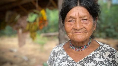 Tsimane woman with dark hair pinned up, wearing a white top with black flowers and a necklace