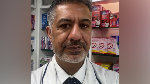 A man with short greying hair and a trimmed beard takes a selfie while wearing a white pharmacist's coat and a dark patterned tie. He stands in front of shelves stocked with boxes of children's medicines including Calpol and paracetamol.