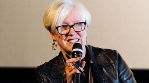 Janice Forsyth in a leather jacket and wearing dark plastic glasses speaking at an event. She is holding a microphone and smiling, her hand waving in the air.