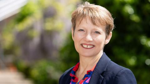 Clare Moody smiles at the camera in a navy blazer and collared patterned shirt. The background is blurred but it appears to be green foliage.