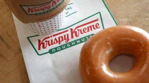 A Krispy Kreme doughnut in a shop. It is glazed with icing and has a hole in the middle. There is also a napkin and a takeaway coffee cup.