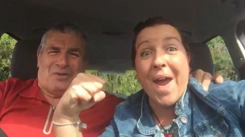 Ali Alcock raises her fist in the air as she and her dad celebrate her being given the all clear. She and her dad are smiling while sitting in his car.